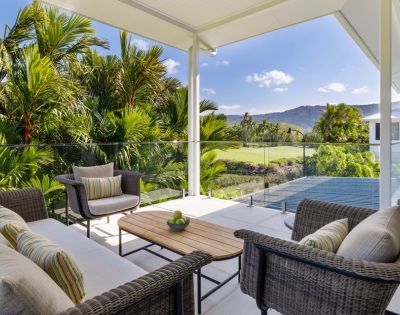 Covered patio with wicker furniture and striped cushions, overlooking lush tropical greenery and distant mountains under a clear blue sky. Relaxing ambiance.