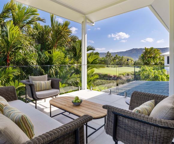 Covered patio with wicker furniture and striped cushions, overlooking lush tropical greenery and distant mountains under a clear blue sky. Relaxing ambiance.