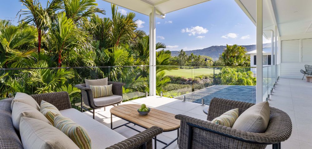 Covered patio with wicker furniture and striped cushions, overlooking lush tropical greenery and distant mountains under a clear blue sky. Relaxing ambiance.