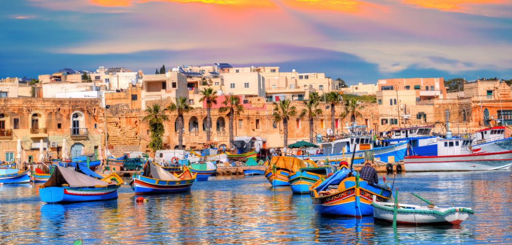 Colorful boats float on a calm harbor with a backdrop of historic buildings and palm trees. The sunset casts warm hues across the sky and water, creating a tranquil atmosphere.