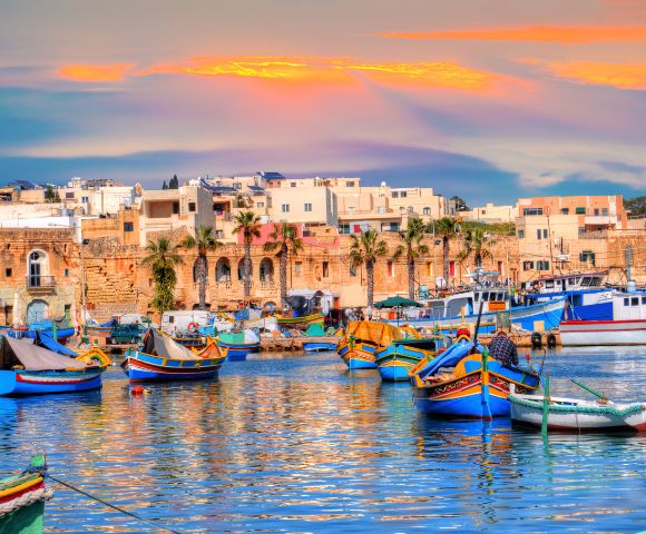 Colorful boats float on a calm harbor with a backdrop of historic buildings and palm trees. The sunset casts warm hues across the sky and water, creating a tranquil atmosphere.