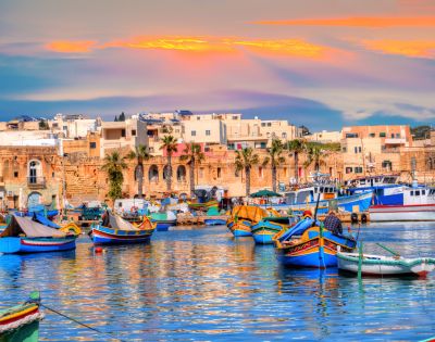Colorful boats float on a calm harbor with a backdrop of historic buildings and palm trees. The sunset casts warm hues across the sky and water, creating a tranquil atmosphere.