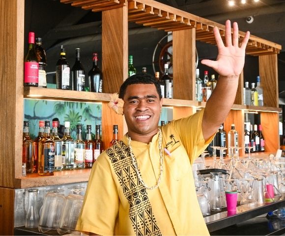 Smiling man with a flower in his hair, wearing a yellow shirt and traditional necklace, waving at the viewer behind a bar stocked with liquor bottles and glasses; wooden shelves and tropical mural background.