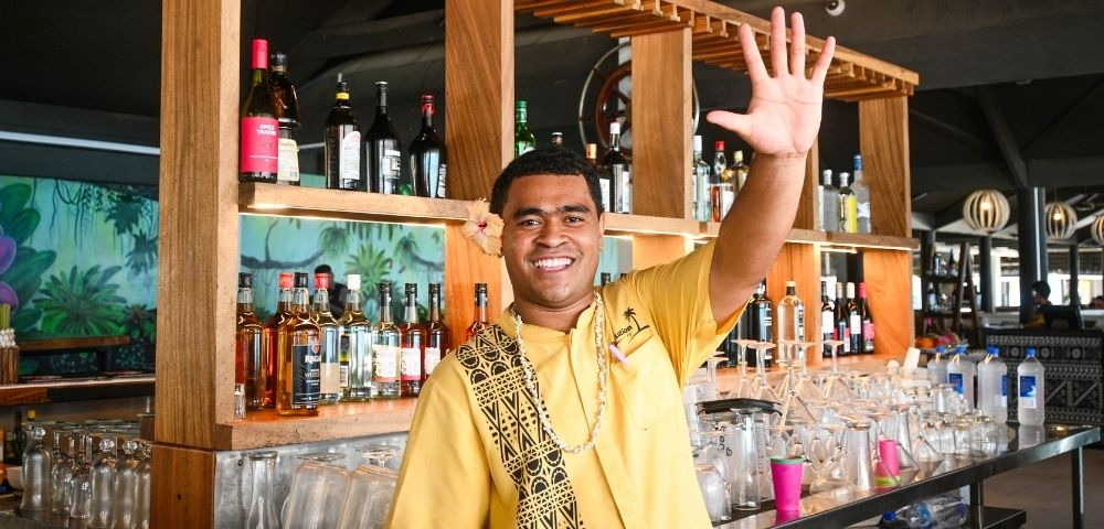 Smiling man with a flower in his hair, wearing a yellow shirt and traditional necklace, waving at the viewer behind a bar stocked with liquor bottles and glasses; wooden shelves and tropical mural background.