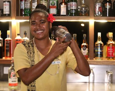 Young woman smiling while holding a cocktail shaker, standing behind a bar with shelves of liquor bottles in the background, wearing a yellow shirt and red flower in her hair.