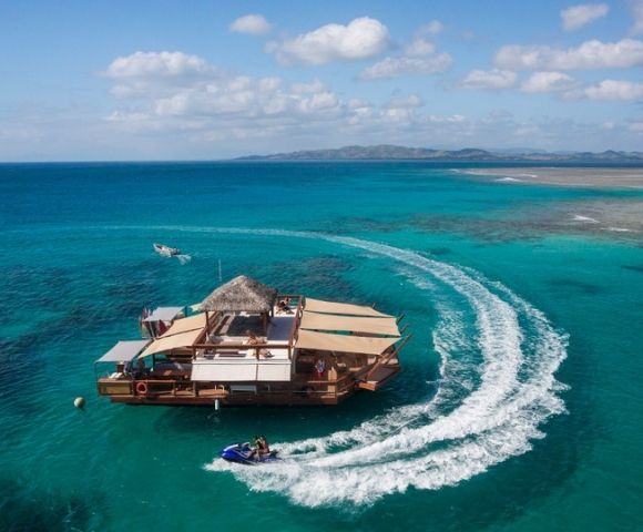 Aerial view of a wooden floating platform with a thatched roof, surrounded by turquoise waters, boats, and distant islands.