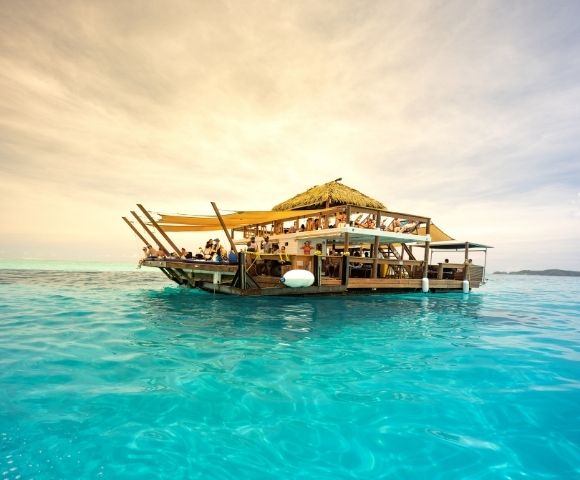 A lively floating bar with a thatched roof, surrounded by turquoise waters, filled with beachgoers enjoying drinks and sun.