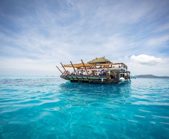 A lively floating restaurant with a thatched roof, surrounded by crystal-clear turquoise waters under a bright sky.