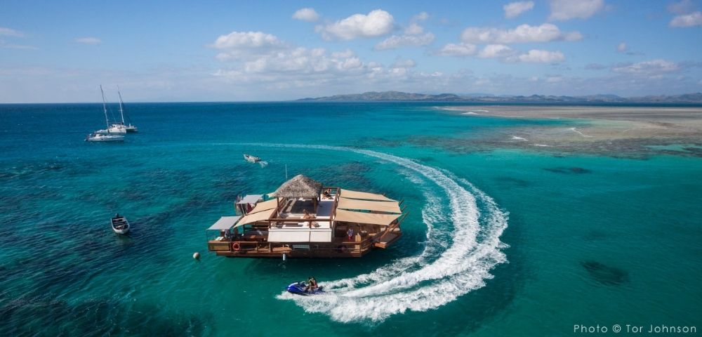 Aerial view of a wooden floating platform with a thatched roof, surrounded by turquoise waters, boats, and distant islands.