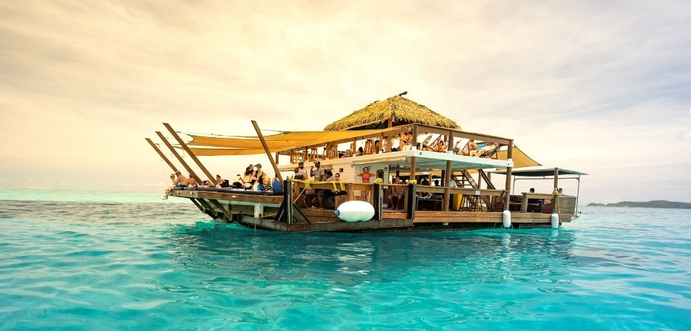 A lively floating bar with a thatched roof, surrounded by turquoise waters, filled with beachgoers enjoying drinks and sun.