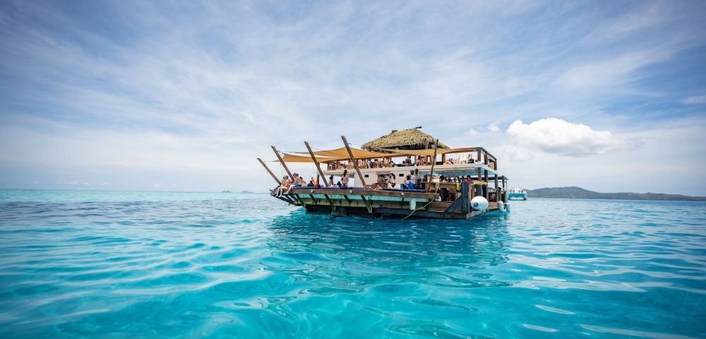 A lively floating restaurant with a thatched roof, surrounded by crystal-clear turquoise waters under a bright sky.
