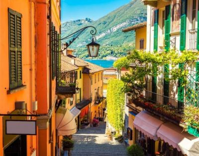 Charming narrow street in an Italian village with colorful buildings, green shutters, and vibrant vines, opening to a serene lake and mountains.