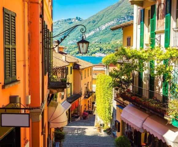 Charming narrow street in an Italian village with colorful buildings, green shutters, and vibrant vines, opening to a serene lake and mountains.
