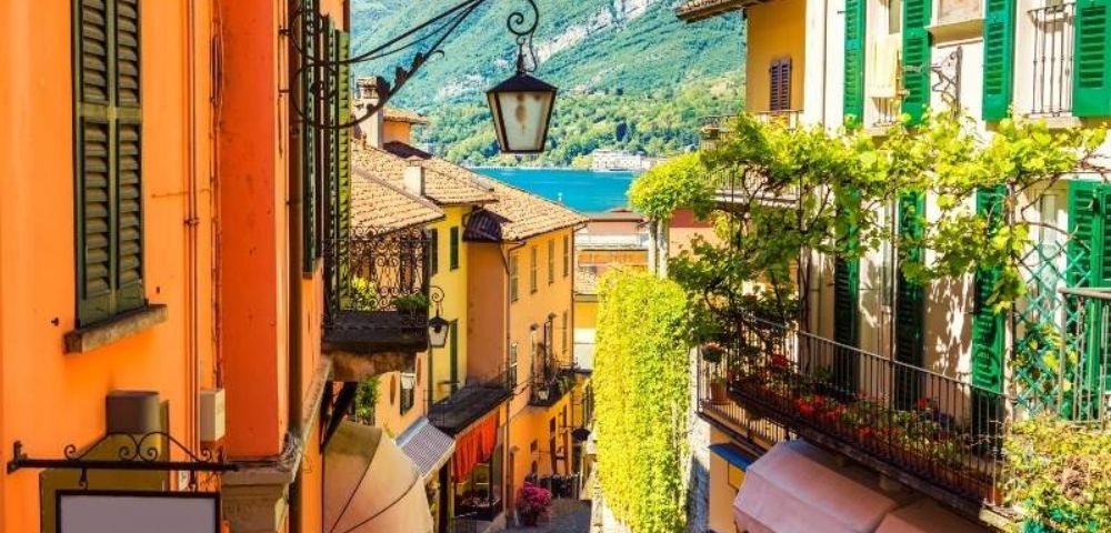 Charming narrow street in an Italian village with colorful buildings, green shutters, and vibrant vines, opening to a serene lake and mountains.