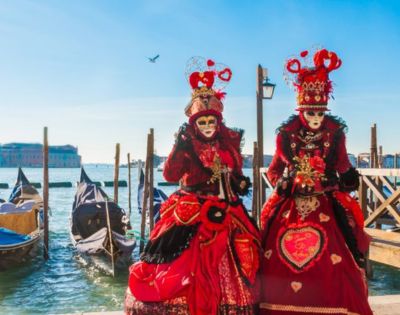 Two figures in ornate red Venetian costumes and masks stand near gondolas by a canal. Bright blue sky and distant buildings enhance a festive atmosphere.