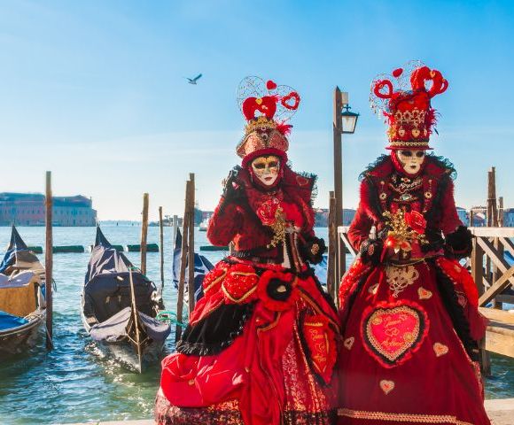 Two figures in ornate red Venetian costumes and masks stand near gondolas by a canal. Bright blue sky and distant buildings enhance a festive atmosphere.