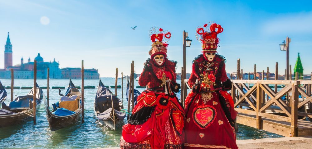 Two figures in ornate red Venetian costumes and masks stand near gondolas by a canal. Bright blue sky and distant buildings enhance a festive atmosphere.