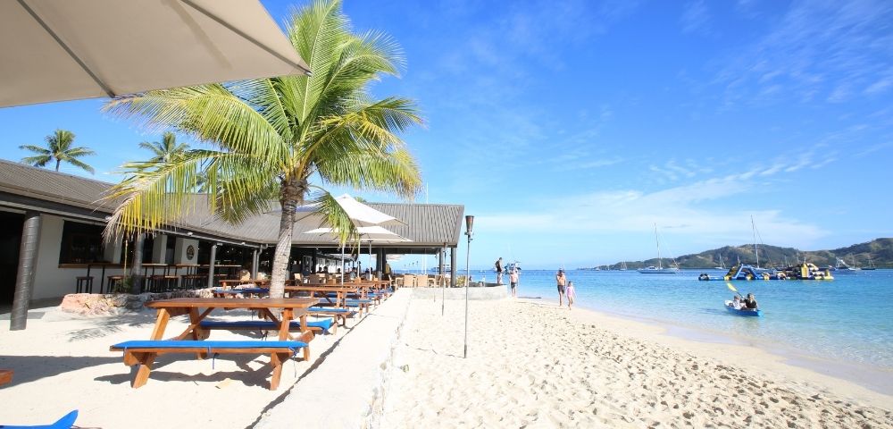 Beachfront restaurant with picnic tables, palm trees, and umbrellas, with a white sand beach and clear blue water visible in the background, people walking on the beach and kayaking.