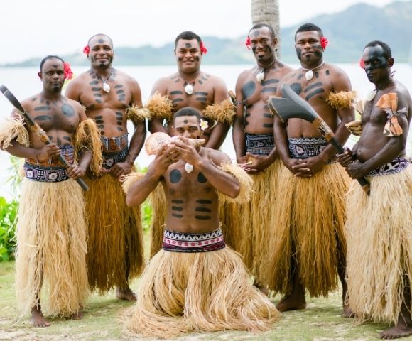 A group of six Fijian men, wearing traditional grass skirts, body paint, and flowers in their hair, posing outdoors with traditional weapons and a conch shell; the men are standing on a grassy area with foliage and water in the background.