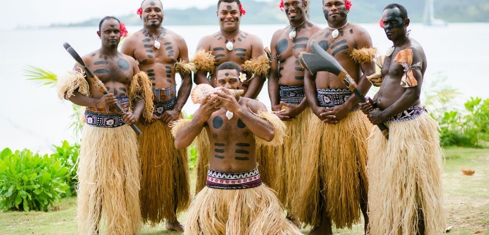 A group of six Fijian men, wearing traditional grass skirts, body paint, and flowers in their hair, posing outdoors with traditional weapons and a conch shell; the men are standing on a grassy area with foliage and water in the background.