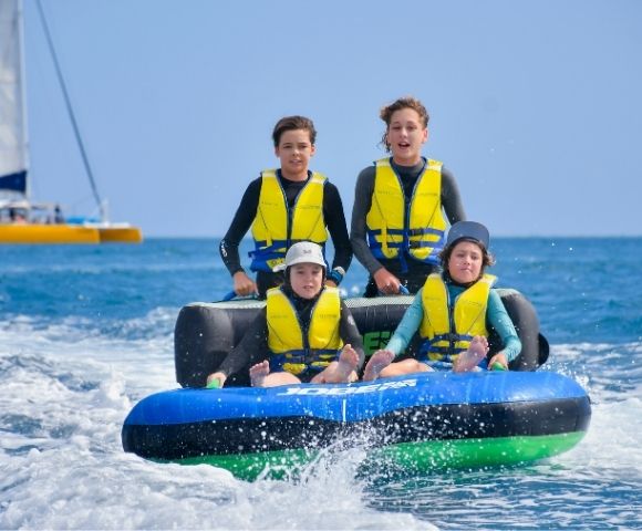 Four children in wetsuits and life vests ride a blue and black inflatable tube on the ocean, with a yellow catamaran in the background and a clear blue sky.