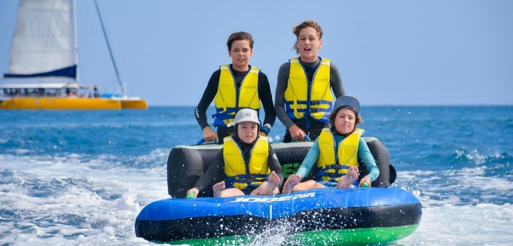 Four children in wetsuits and life vests ride a blue and black inflatable tube on the ocean, with a yellow catamaran in the background and a clear blue sky.