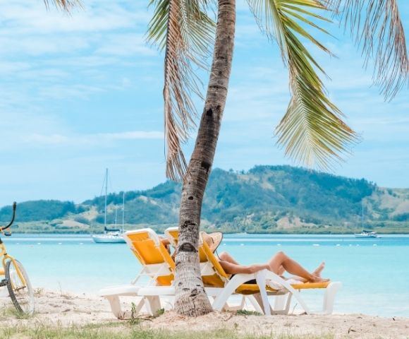 Person relaxing in a yellow beach chair under a palm tree on a tropical beach, with a yellow bicycle, blue water, distant sailboats, and mountains in the background.