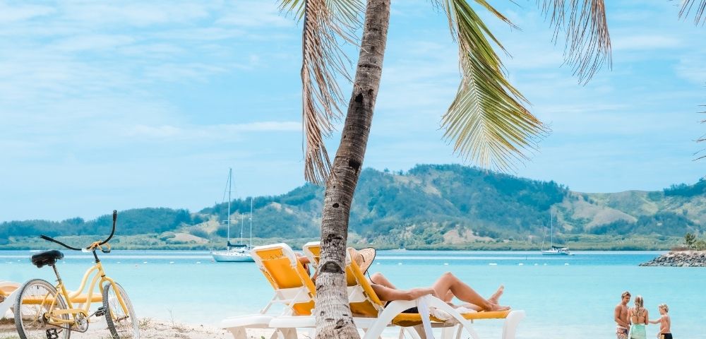 Person relaxing in a yellow beach chair under a palm tree on a tropical beach, with a yellow bicycle, blue water, distant sailboats, and mountains in the background.