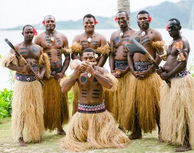 A group of six Fijian men, wearing traditional grass skirts, body paint, and flowers in their hair, posing outdoors with traditional weapons and a conch shell; the men are standing on a grassy area with foliage and water in the background.