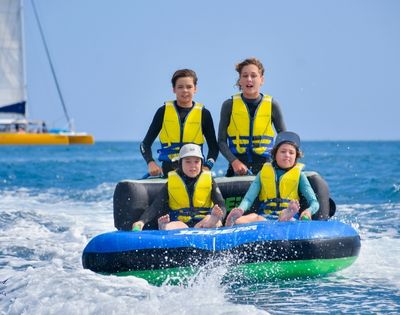Four children in wetsuits and life vests ride a blue and black inflatable tube on the ocean, with a yellow catamaran in the background and a clear blue sky.