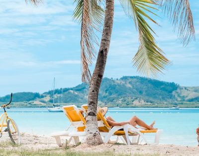 Person relaxing in a yellow beach chair under a palm tree on a tropical beach, with a yellow bicycle, blue water, distant sailboats, and mountains in the background.