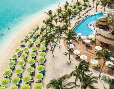 Aerial view of a vibrant beach resort with turquoise water, green umbrellas, and sunbeds on white sand. A winding pool is fringed by palm trees and a wooden deck bar.