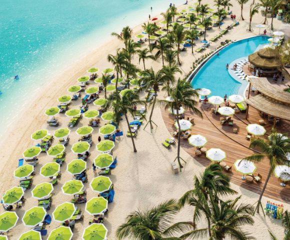 Aerial view of a vibrant beach resort with turquoise water, green umbrellas, and sunbeds on white sand. A winding pool is fringed by palm trees and a wooden deck bar.