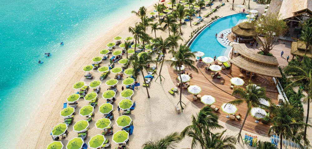 Aerial view of a vibrant beach resort with turquoise water, green umbrellas, and sunbeds on white sand. A winding pool is fringed by palm trees and a wooden deck bar.