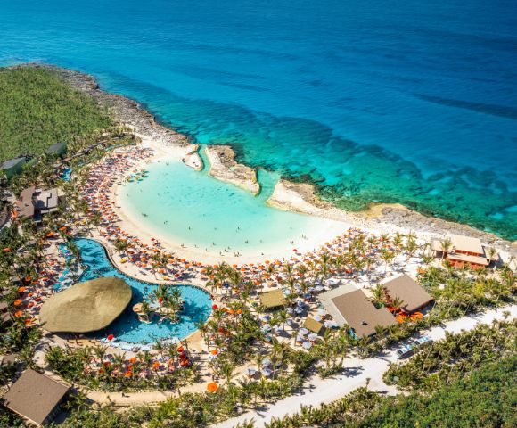Aerial view of a tropical beach resort with turquoise ocean, sandy beach, and a pool surrounded by palm trees and colorful umbrellas, exuding a vibrant, relaxing atmosphere.