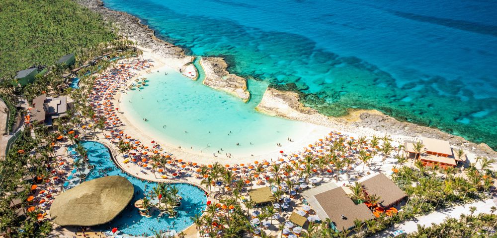 Aerial view of a tropical beach resort with turquoise ocean, sandy beach, and a pool surrounded by palm trees and colorful umbrellas, exuding a vibrant, relaxing atmosphere.