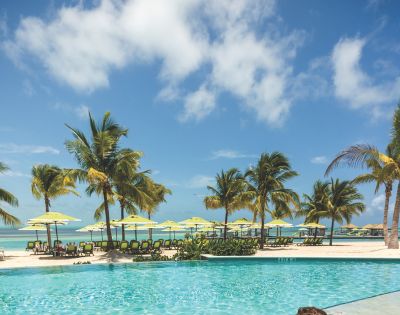 Tropical beach scene with palm trees, yellow beach umbrellas, and lounge chairs on white sand. A bright blue sky and turquoise ocean create a relaxing vibe.