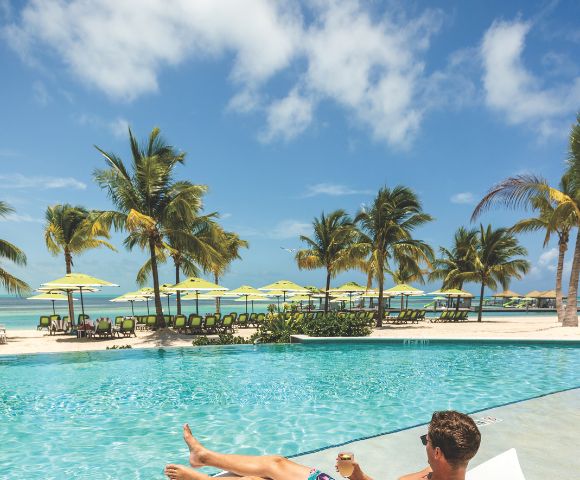 Tropical beach scene with palm trees, yellow beach umbrellas, and lounge chairs on white sand. A bright blue sky and turquoise ocean create a relaxing vibe.