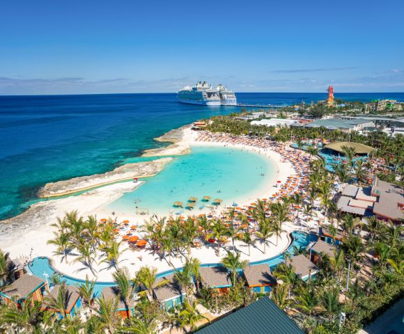 A vibrant beach scene with turquoise water, white sand, and colorful umbrellas. A cruise ship is docked nearby, surrounded by lush palm trees and clear skies.
