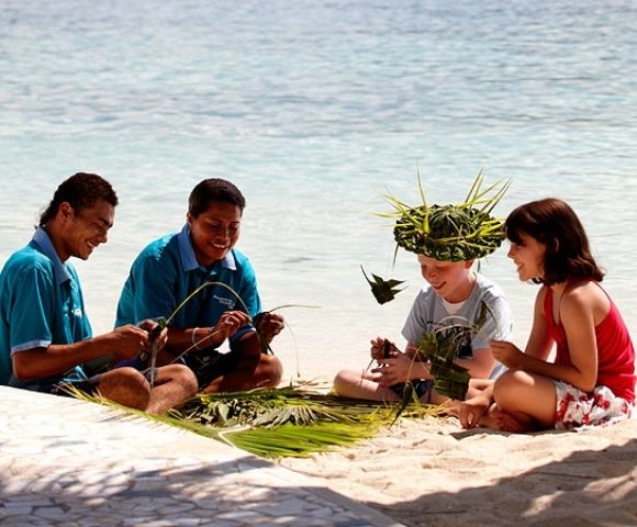 Four people sitting on a sandy beach with the ocean in the background. Two men in blue shirts, and two children are making crafts from green leaves, with one child wearing a leaf hat. A palm tree trunk is on the right side of the image.
