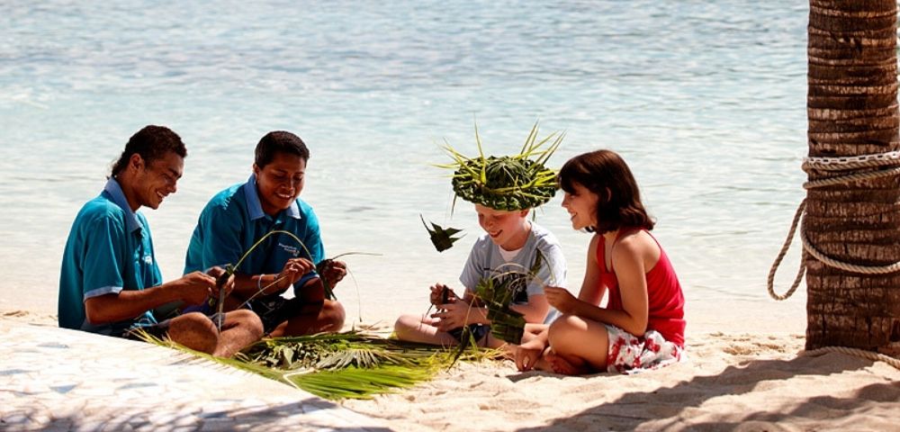 Four people sitting on a sandy beach with the ocean in the background. Two men in blue shirts, and two children are making crafts from green leaves, with one child wearing a leaf hat. A palm tree trunk is on the right side of the image.