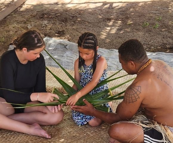 Three people weaving palm fronds together while sitting on a woven mat. The person on the right, a man, is shirtless with a thick gold necklace and tattoos on his back. Two young girls are on the left and center wearing a black shirt and floral sundress, respectively. They're all focused on manipulating the leaves.