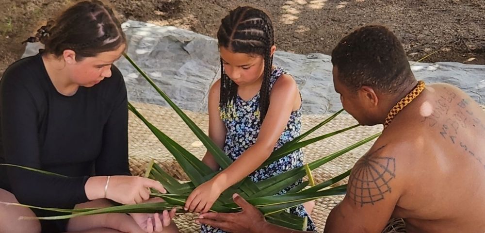 Three people weaving palm fronds together while sitting on a woven mat. The person on the right, a man, is shirtless with a thick gold necklace and tattoos on his back. Two young girls are on the left and center wearing a black shirt and floral sundress, respectively. They're all focused on manipulating the leaves.