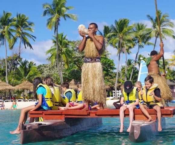 A Polynesian man in traditional attire blows into a conch shell while standing on a watercraft, surrounded by children wearing life vests, with a tropical beach scene in the background under a bright blue sky.