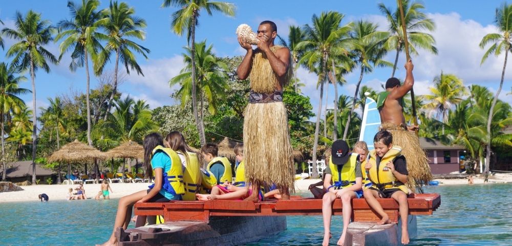 A Polynesian man in traditional attire blows into a conch shell while standing on a watercraft, surrounded by children wearing life vests, with a tropical beach scene in the background under a bright blue sky.