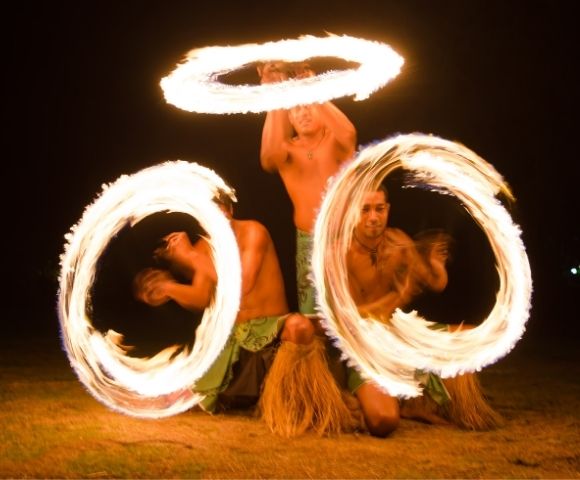 Three shirtless men performing a fire dance at night, each spinning flaming hoops around themselves and in the air, the flames creating light streaks.