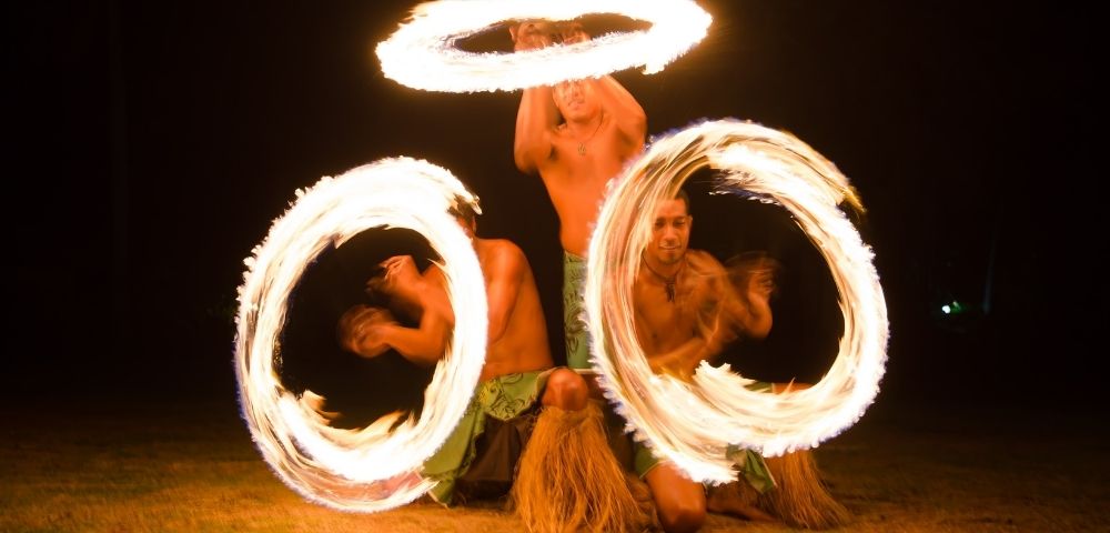 Three shirtless men performing a fire dance at night, each spinning flaming hoops around themselves and in the air, the flames creating light streaks.