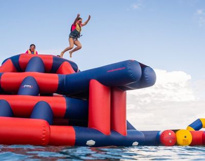 A girl in a life vest jumping into the air off a large, inflatable water park obstacle course, with another girl wearing glasses in the background on top of the obstacle course.
