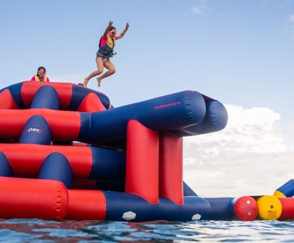 A girl in a life vest jumping into the air off a large, inflatable water park obstacle course, with another girl wearing glasses in the background on top of the obstacle course.