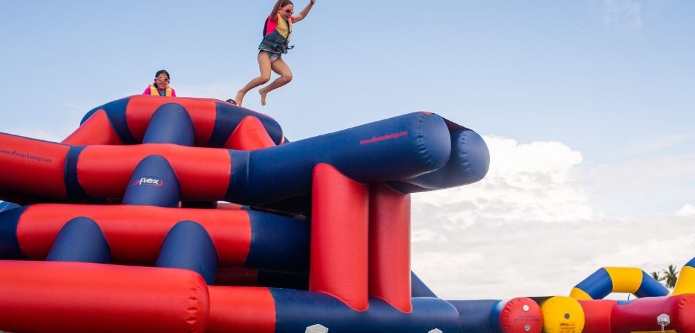 A girl in a life vest jumping into the air off a large, inflatable water park obstacle course, with another girl wearing glasses in the background on top of the obstacle course.